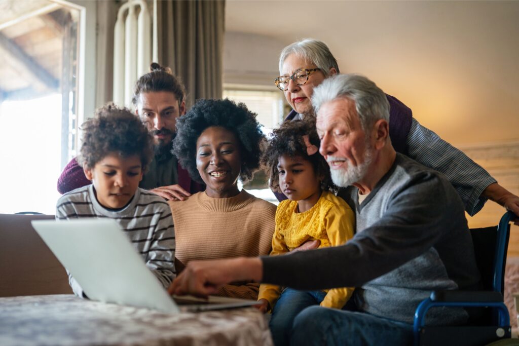 Happy multi generational and multiracial family sitting together at dinner table looking at laptop.